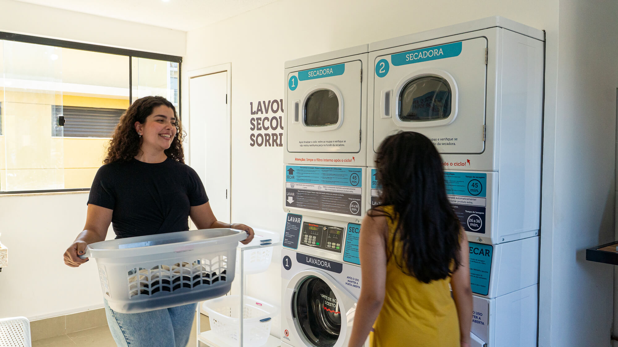 Moradora com cesto de roupas diante das lavadoras e secadoras da lavanderia autônoma da Roda em um condomínio